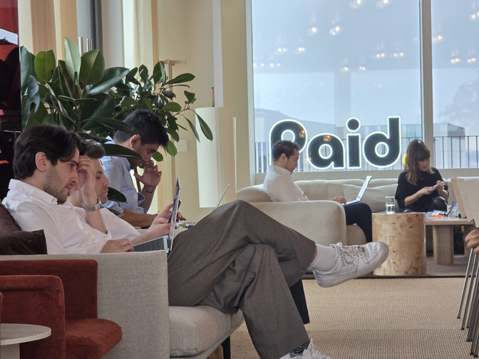 People sitting on couches in a modern lounge, using laptops and phones. Large windows display "Paid" in the background.