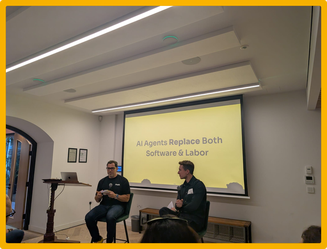 Two people sit on stools in front of a screen displaying "AI Agents Replace Both Software & Labor" during a presentation.